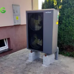 Szara pompa ciepła marki Nesheat, zamontowana na betonowych bloczkach przed budynkiem z oknem piwnicznym i skrzynką elektryczną na elewacji.