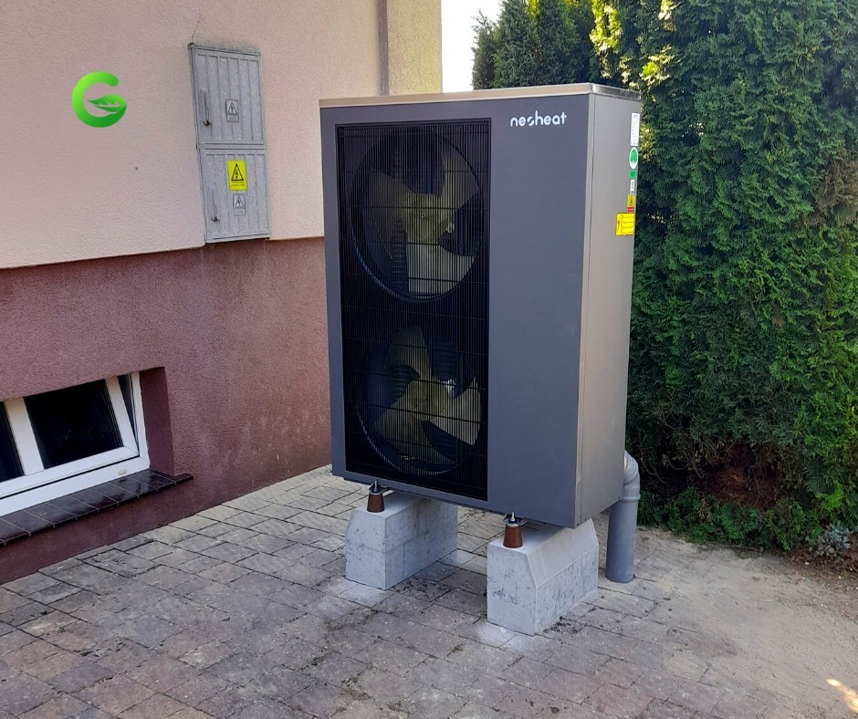 Szara pompa ciepła marki Nesheat, zamontowana na betonowych bloczkach przed budynkiem z oknem piwnicznym i skrzynką elektryczną na elewacji.