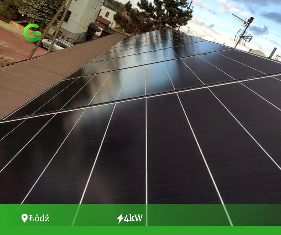 Panele słoneczne na brązowym dachu budynku mieszkalnego, widoczne anteny telewizyjne i odbicie nieba w panelach.