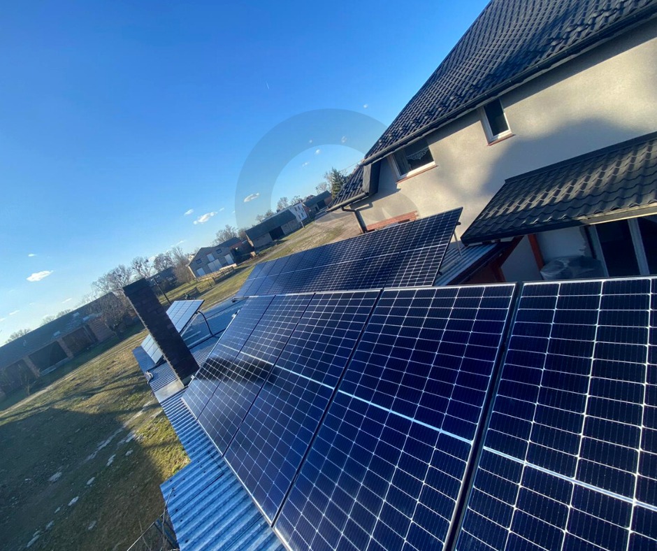 Panele słoneczne na dachu domu z szarej elewacją i czarną dachówką, widok z perspektywy dachu, błękitne niebo w tle.