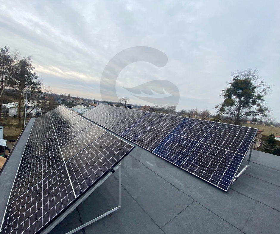 Instalacja paneli słonecznych na płaskim dachu pokrytym papą, widok z góry na tle pochmurnego nieba i zabudowań mieszkalnych.