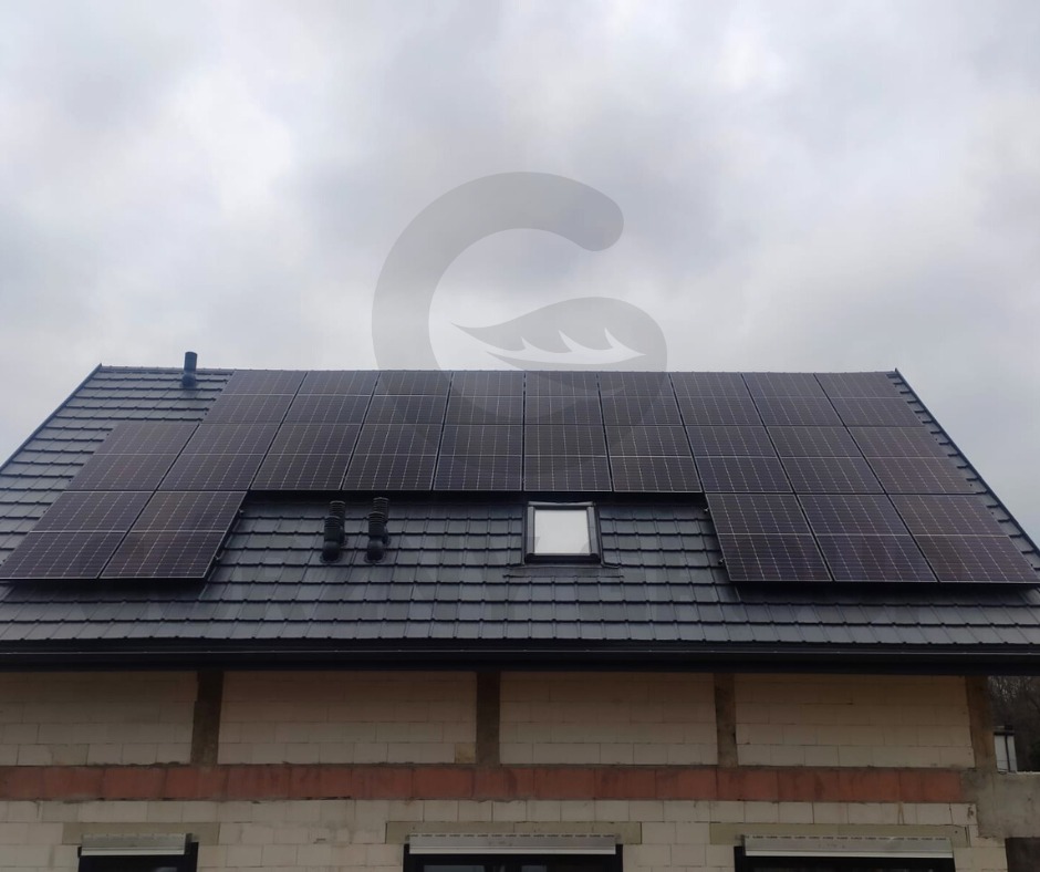 Panele słoneczne na dachu domu z ciemnej dachówki, widok z dołu na pochmurnym tle.