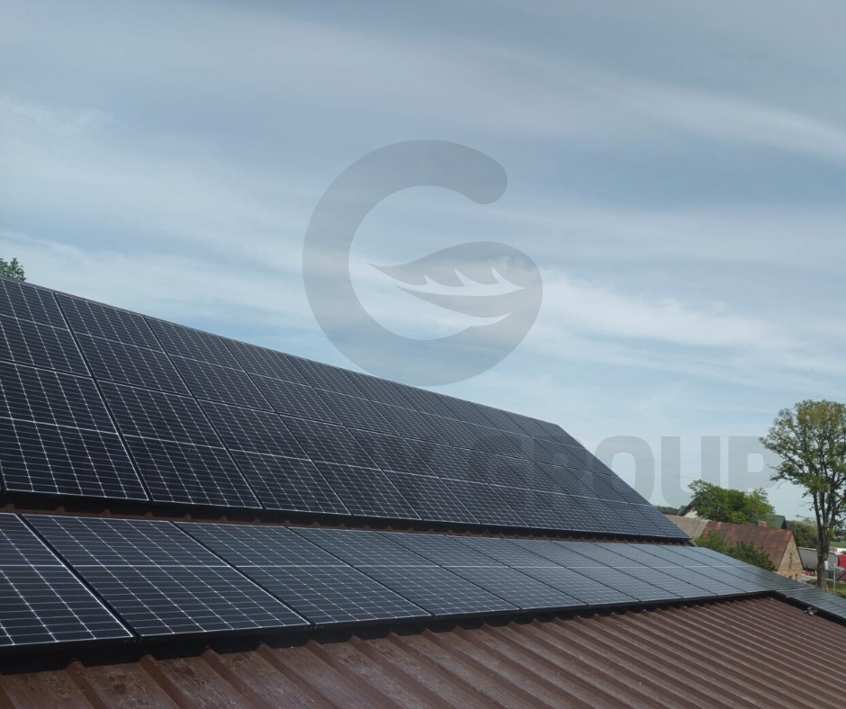 Panele fotowoltaiczne zamontowane na dachu z blachy trapezowej w wiejskiej scenerii, widoczne drzewa i budynki w tle.