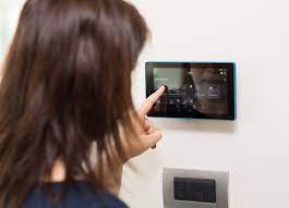 Kobieta obsługuje dotykowy panel sterowania systemu inteligentnego domu zamontowany na ścianie, widoczne ikony i interfejs użytkownika.