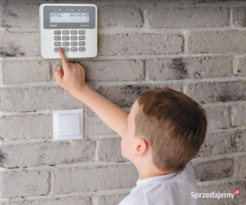 Chłopiec wciska przycisk na panelu alarmowym Satel Perfecto, zamontowanym na ścianie z cegły, obok włącznika światła.