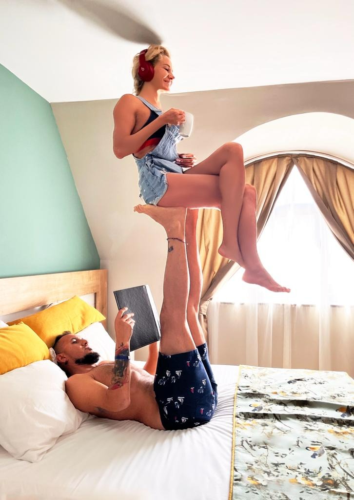 Mężczyzna leżący na łóżku czyta książkę, podczas gdy kobieta w słuchawkach siedzi na jego stopach, trzymając kubek, tworząc akrobatyczną pozę w jasnym pokoju.