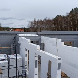 Budowa Domów Pasywnych 17 - Wznoszenie ścian budynku z białych i szarych bloczków styropianowych, widoczne paczki materiałów budowlanych i rusztowanie, w tle drzewa i pochmurne niebo.