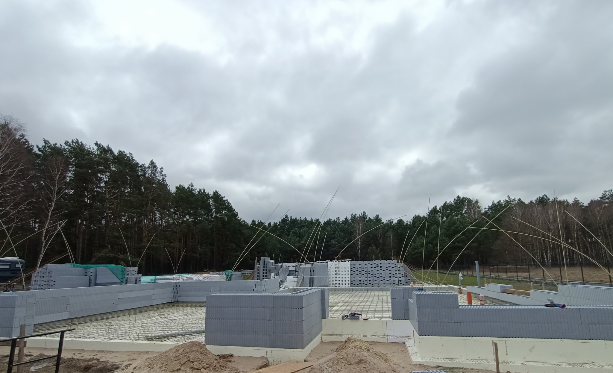 Budowa domu w stanie surowym otwartym, widoczne fundamenty, ściany z bloczków betonowych, zbrojenie, teren budowy, w tle las i pochmurne niebo.