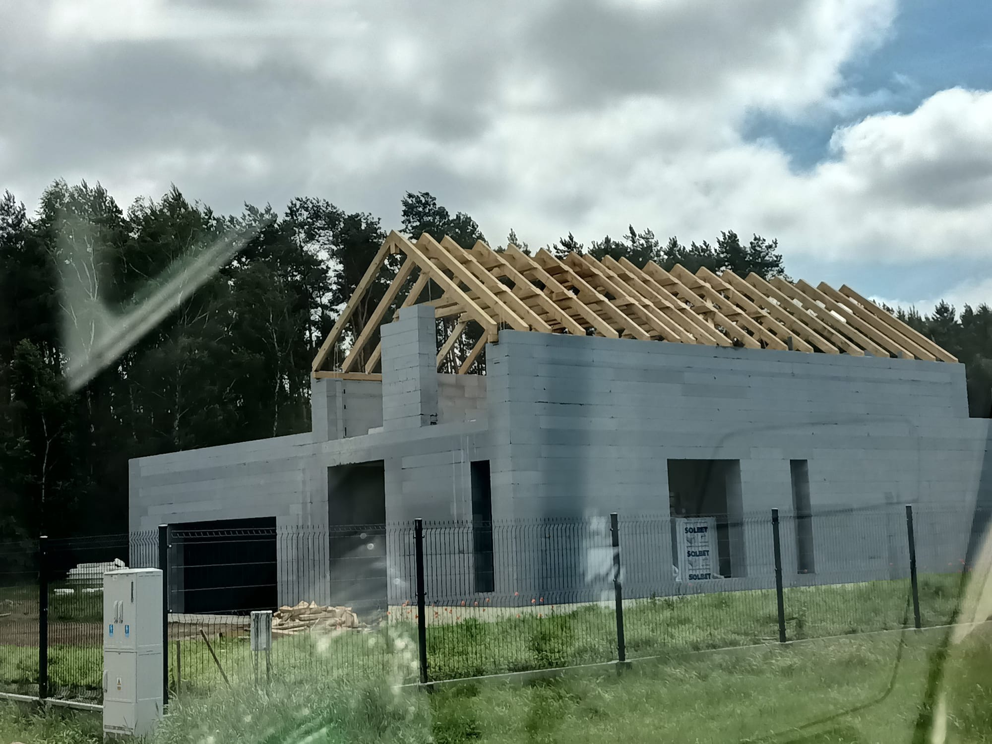 Budowa domu jednorodzinnego w stanie surowym otwartym, ściany z szarego betonu komórkowego, drewniana konstrukcja dachu, widok zza ogrodzenia.