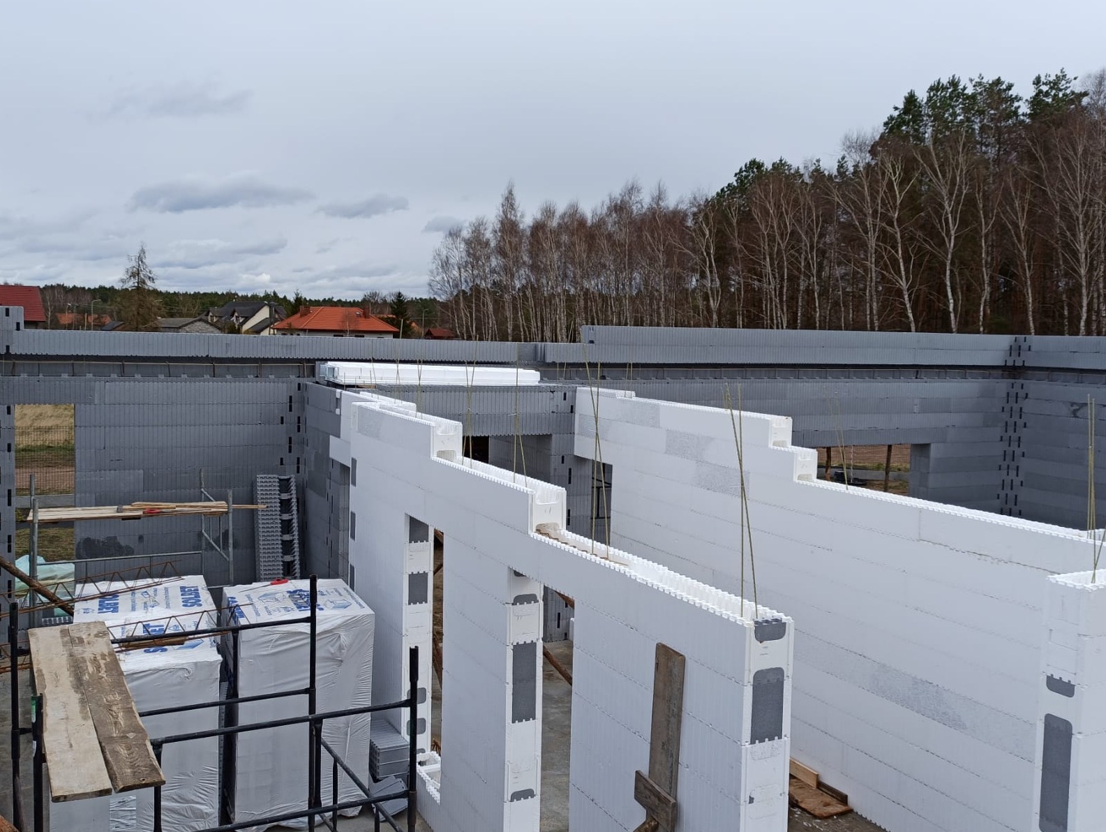 Wznoszenie ścian budynku z białych i szarych bloczków styropianowych, widoczne paczki materiałów budowlanych i rusztowanie, w tle drzewa i pochmurne niebo.