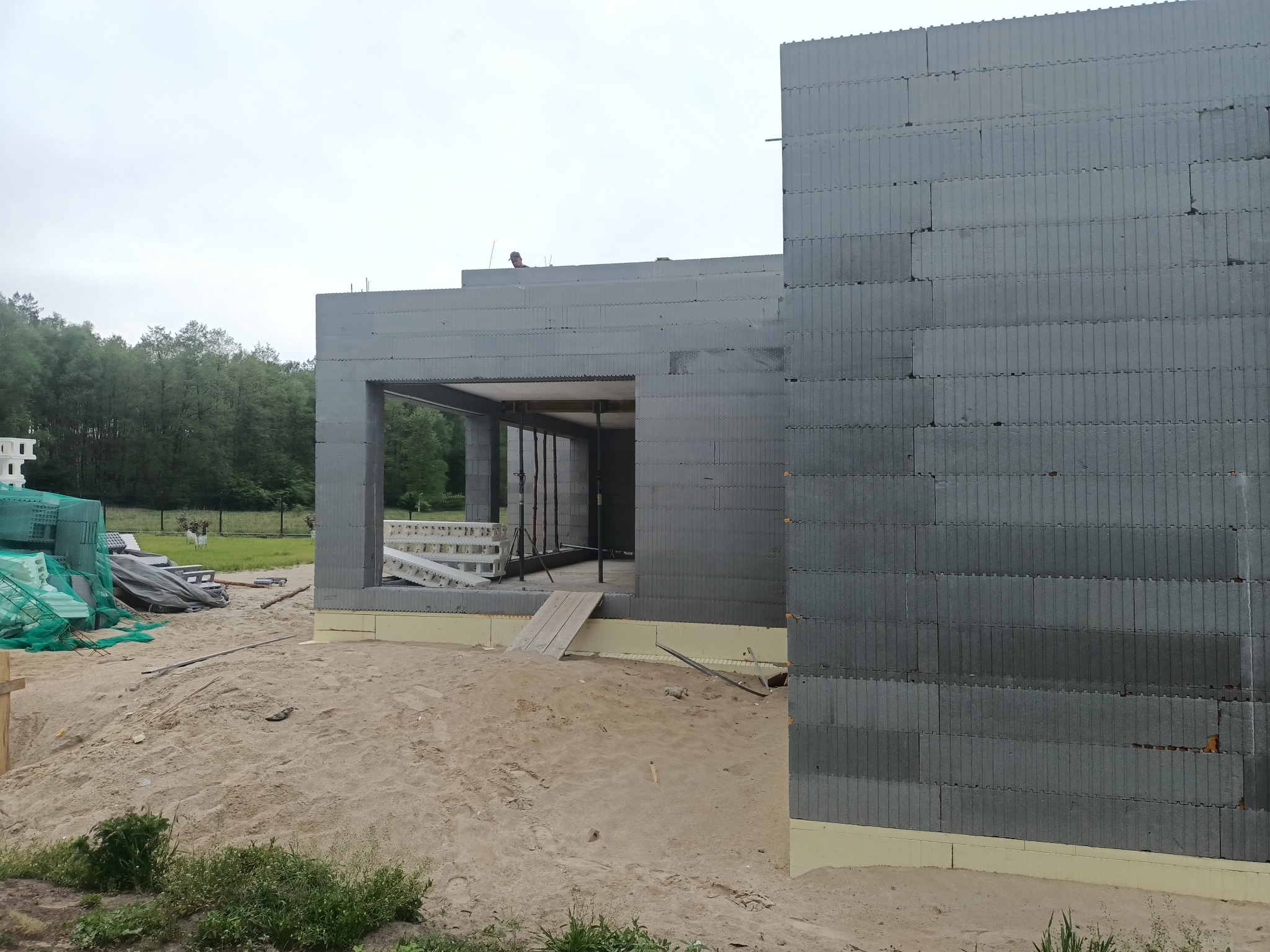 Budowa domu w stanie surowym otwartym z bloczków betonowych, widoczny fragment ściany z oknem, piasek na gruncie, w tle las.