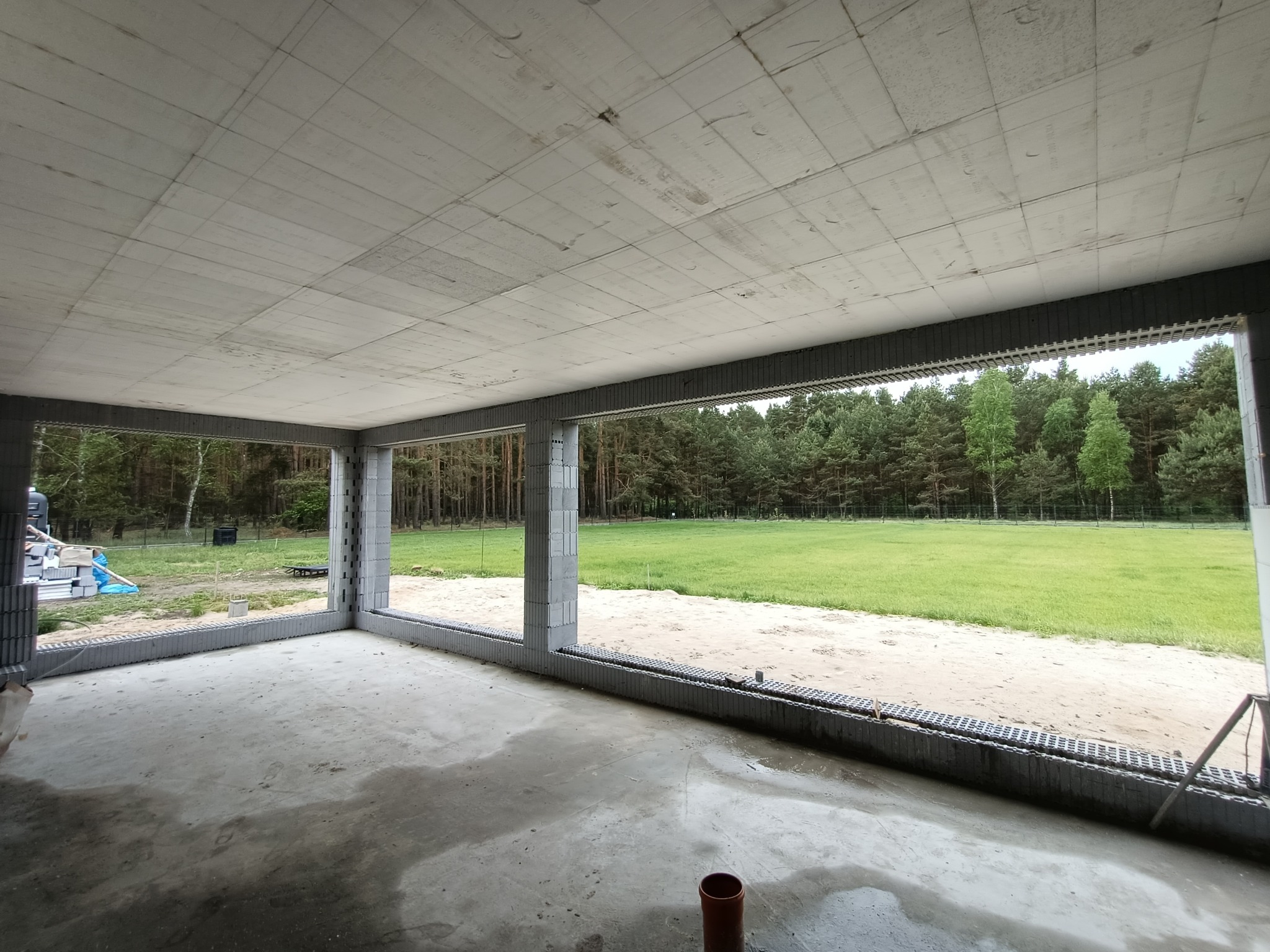 Surowy stan wnętrza budynku w trakcie budowy, widoczne ściany z betonu komórkowego, surowy betonowy sufit i wylewka podłogowa, z widokiem na zewnątrz na łąkę i las.