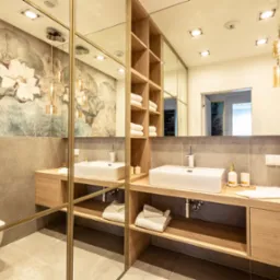 Nowoczesna łazienka z dwoma umywalkami na drewnianych blatach, duże lustro z złotymi ramami, na ścianie tapeta z motywem kwiatowym, wisząca lampa z bursztynowego szkła.