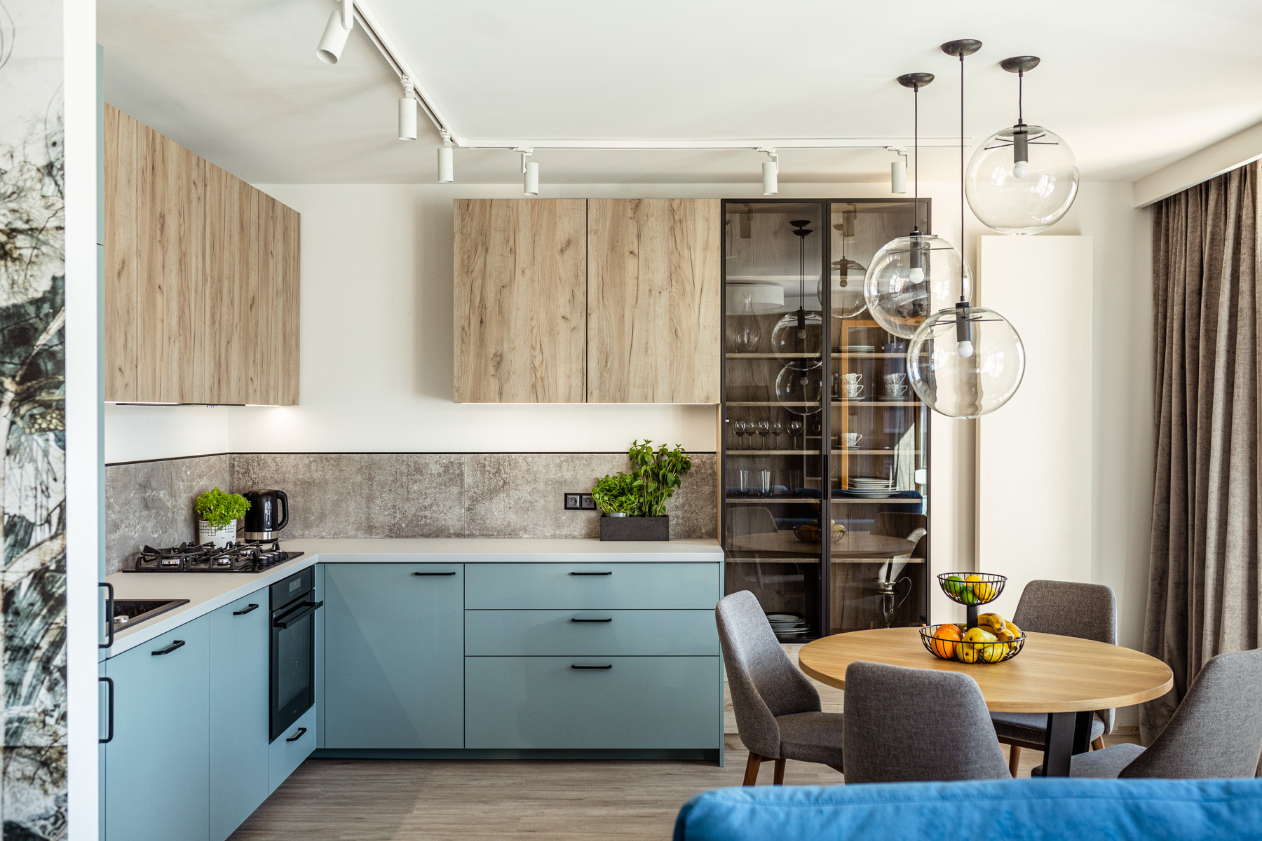 Jasna kuchnia z niebieskimi szafkami, drewnianymi górnymi szafkami, okrągłym stołem i szklaną witryną z zastawą, oświetlona trzema wiszącymi lampami kulami.