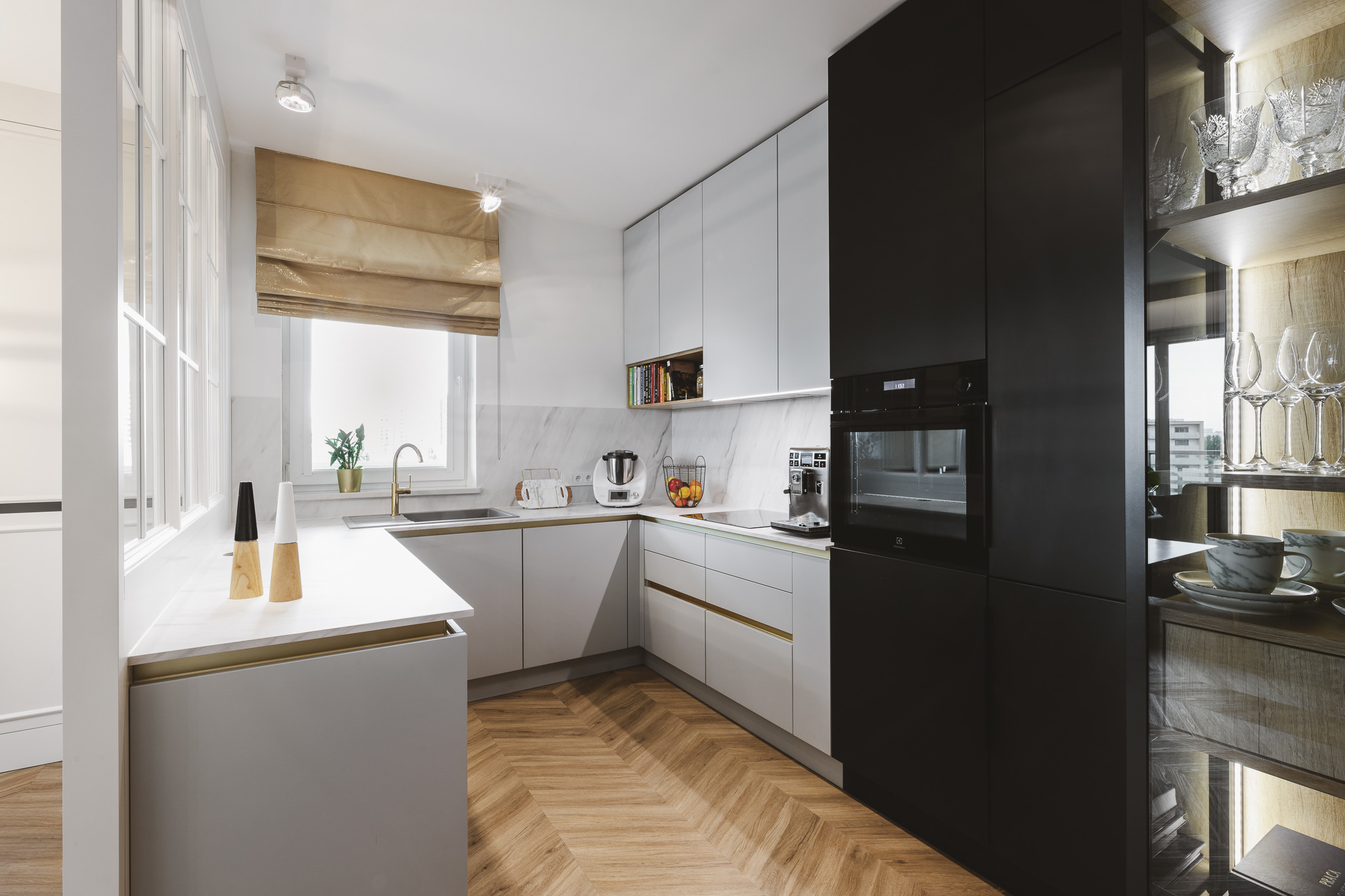 Nowoczesna kuchnia w jasnych szarościach z akcentami czerni i złotymi detalami, parkiet ułożony w jodełkę, przeszklona witryna z kieliszkami.