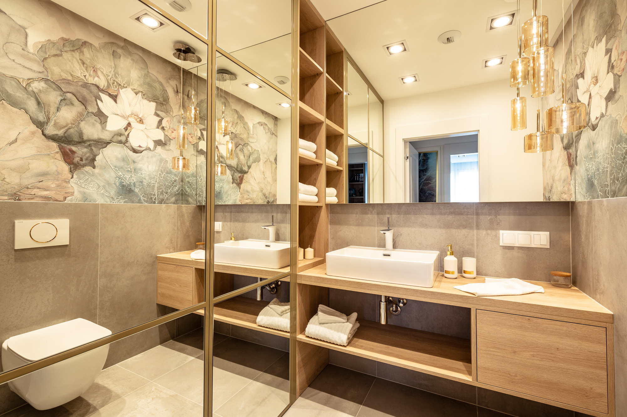 Nowoczesna łazienka z dwoma umywalkami na drewnianych blatach, duże lustro z złotymi ramami, na ścianie tapeta z motywem kwiatowym, wisząca lampa z bursztynowego szkła.