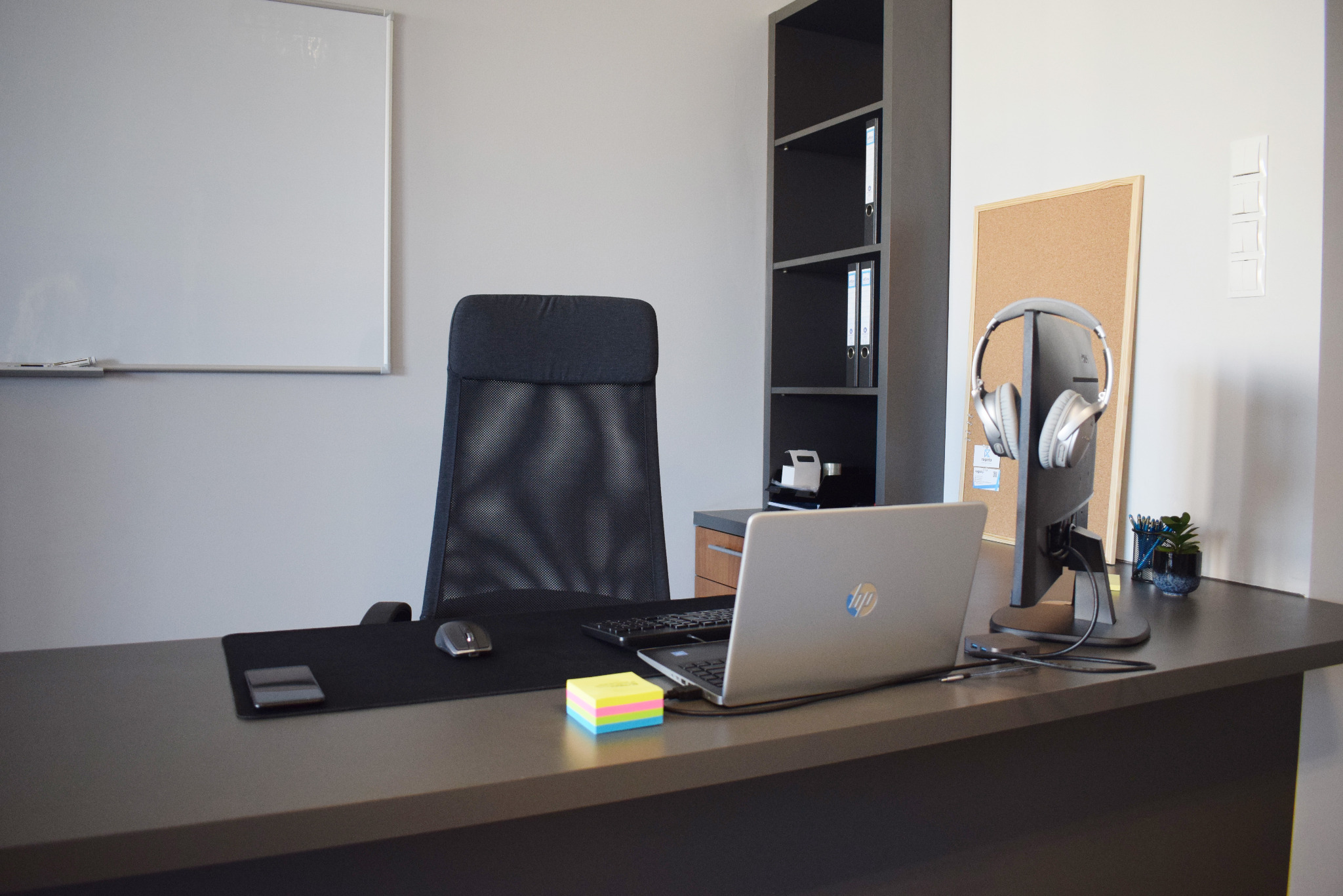 Nowoczesne biuro z ergonomicznym krzesłem, laptopem HP, monitorem z słuchawkami, biurkiem w kolorze grafitowym, karteczkami samoprzylepnymi i tablicą suchościeralną w tle.