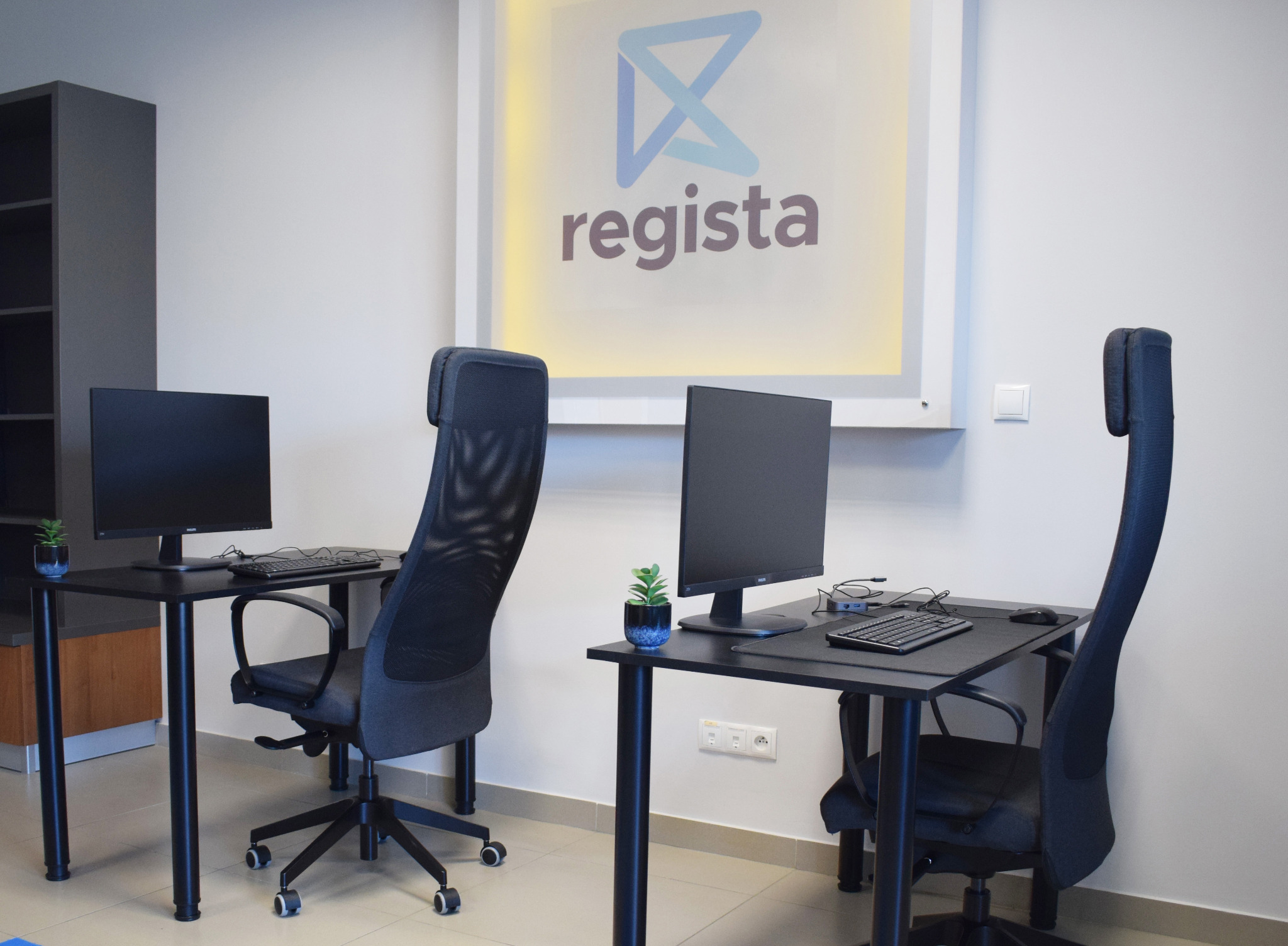 Nowoczesne biuro z dwoma stanowiskami pracy: czarne biurka z komputerami, ergonomiczne krzesła z siatki i dekoracyjne rośliny w doniczkach, logo firmy Regista podświetlone na ścianie.