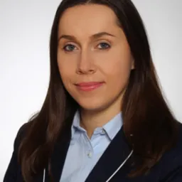 Portret kobiety w niebieskiej koszuli i granatowej marynarce z białą lamówką, uśmiechającej się do kamery na jasnym tle.