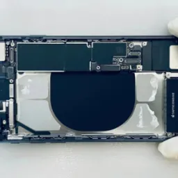 Wnętrze rozłożonego smartfona iPhone z widoczną baterią, elementami elektroniki i silnikiem Taptic Engine, trzymane w dłoniach w białych rękawiczkach ochronnych na jasnym tle.