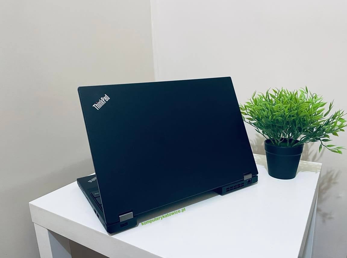 Czarny laptop ThinkPad z widocznym logo, ustawiony na białym stoliku obok zielonej rośliny doniczkowej, widok z tyłu obudowy.