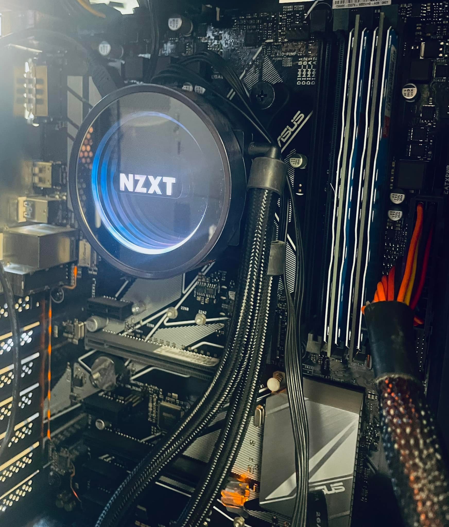 Wnętrze komputera stacjonarnego z widocznym chłodzeniem wodnym procesora marki NZXT, płytą główną ASUS, modułami RAM z podświetleniem LED i okablowaniem zasilającym.