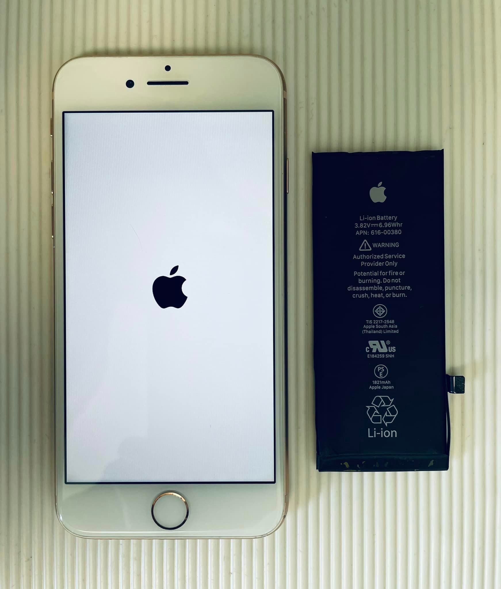 Złoty iPhone z włączonym ekranem prezentującym logo Apple obok czarnej, oryginalnej baterii litowo-jonowej iPhone'a, z widocznymi specyfikacjami technicznymi i ostrzeżeniami.