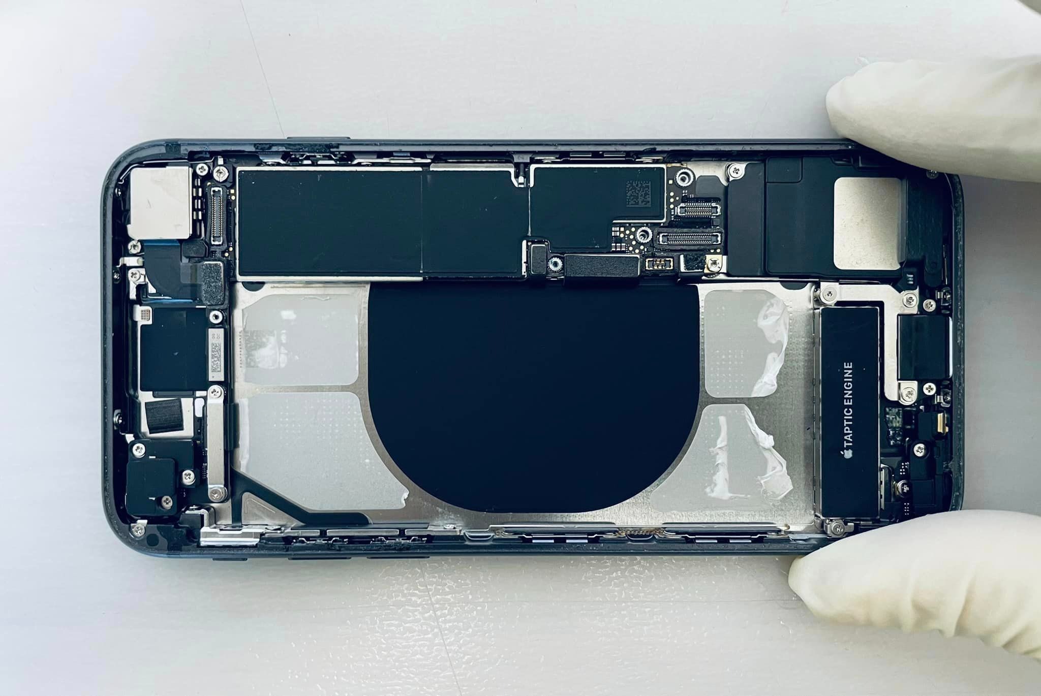 Wnętrze rozłożonego smartfona iPhone z widoczną baterią, elementami elektroniki i silnikiem Taptic Engine, trzymane w dłoniach w białych rękawiczkach ochronnych na jasnym tle.