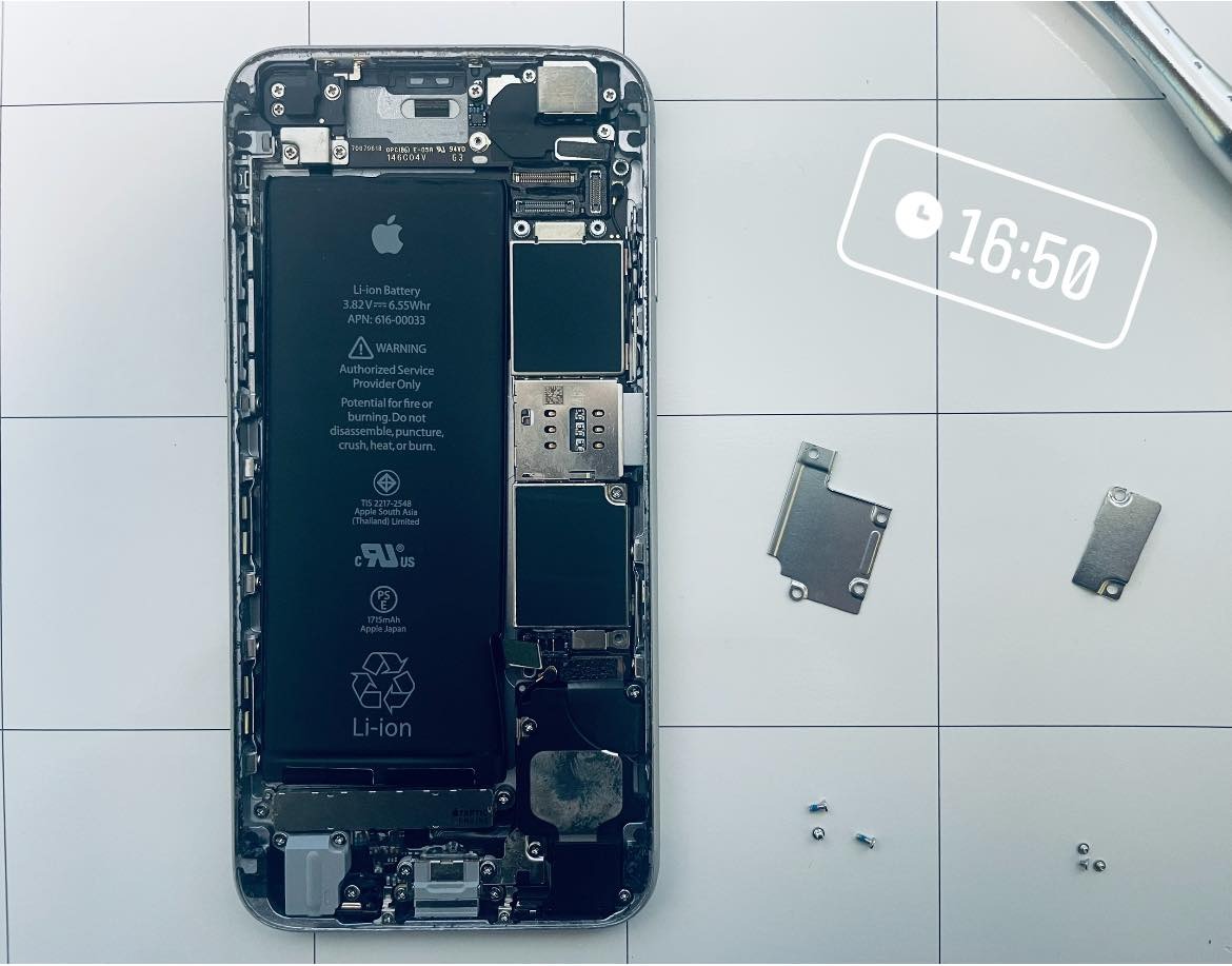 Wnętrze obudowy rozłożonego iPhone'a z widoczną baterią, płytą główną i elementami mocującymi na białym, kwadratowym podłożu. Obok leżą metalowe osłonki i małe śrubki.
