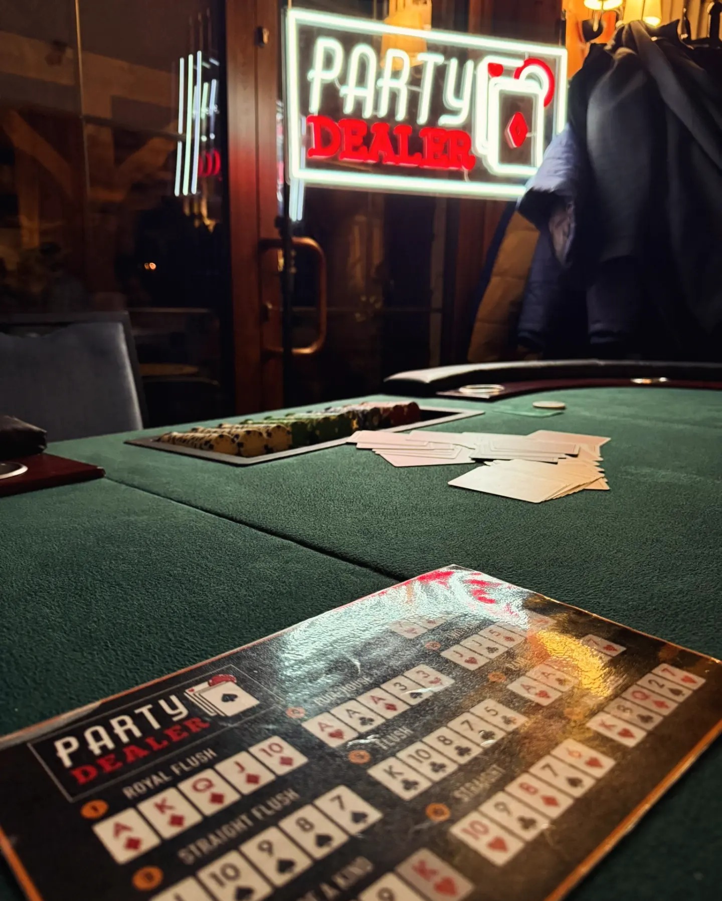 Stół do pokera z żetonami, kartami i neonem 'Party Dealer' w tle. Widoczne zasady gry. Klimatyczne oświetlenie tworzy intymną atmosferę.