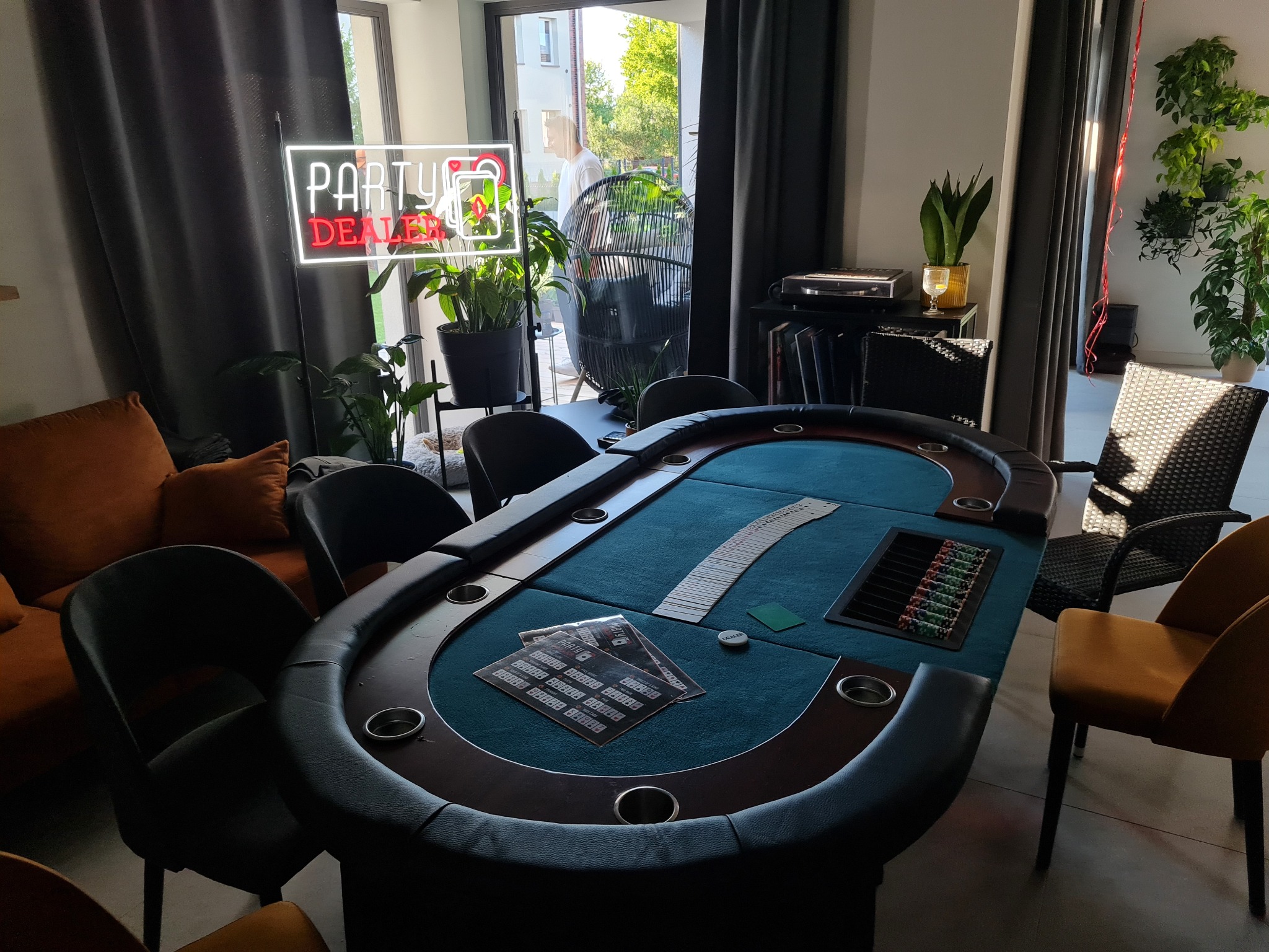 Stół do pokera z kartami i żetonami w eleganckim wnętrzu. Neon 'Party Dealer' i rośliny w tle dodają klimatu.