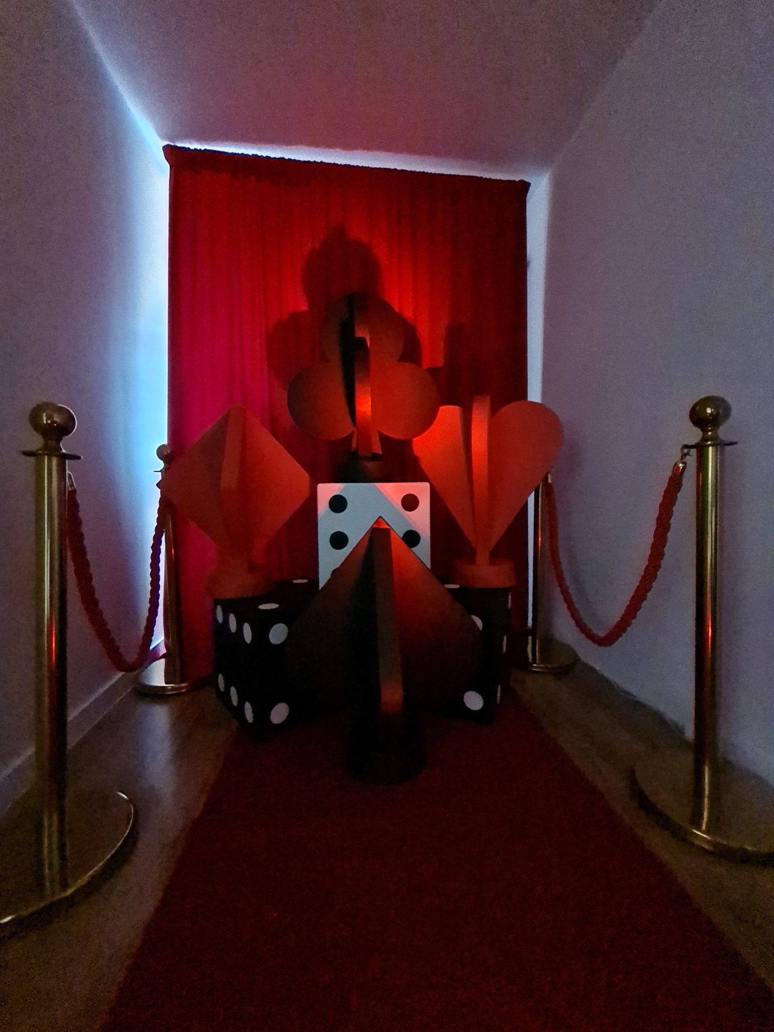 Scena z czerwonym dywanem i dekoracjami karcianymi (karo, trefl, kier, pik) oraz kostkami, otoczona złotymi słupkami z czerwonym sznurem, tworząca iluzję wejścia do kasyna.
