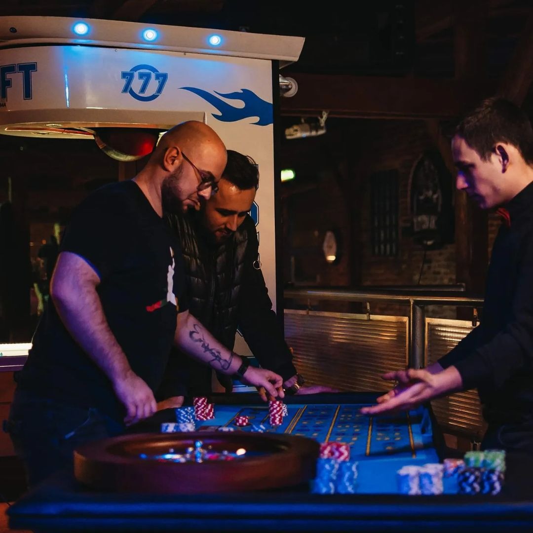 Trzej mężczyźni przy stole do ruletki z żetonami, w tle oświetlony bar z logo 777, ciemne wnętrze lokalu.