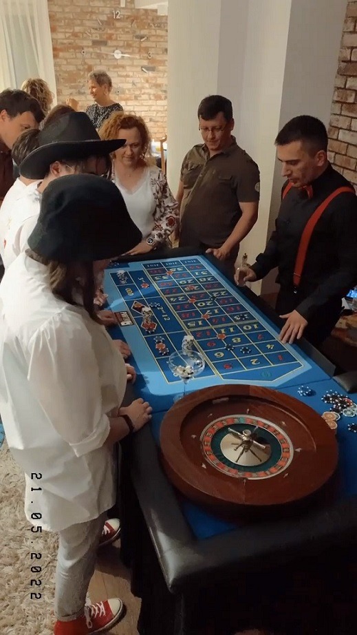 Stół do ruletki z obstawiającymi graczami podczas imprezy w Katowicach. Krupier w czarnej koszuli z czerwonymi szelkami nadzoruje grę. Widoczna data 21.05.2022.