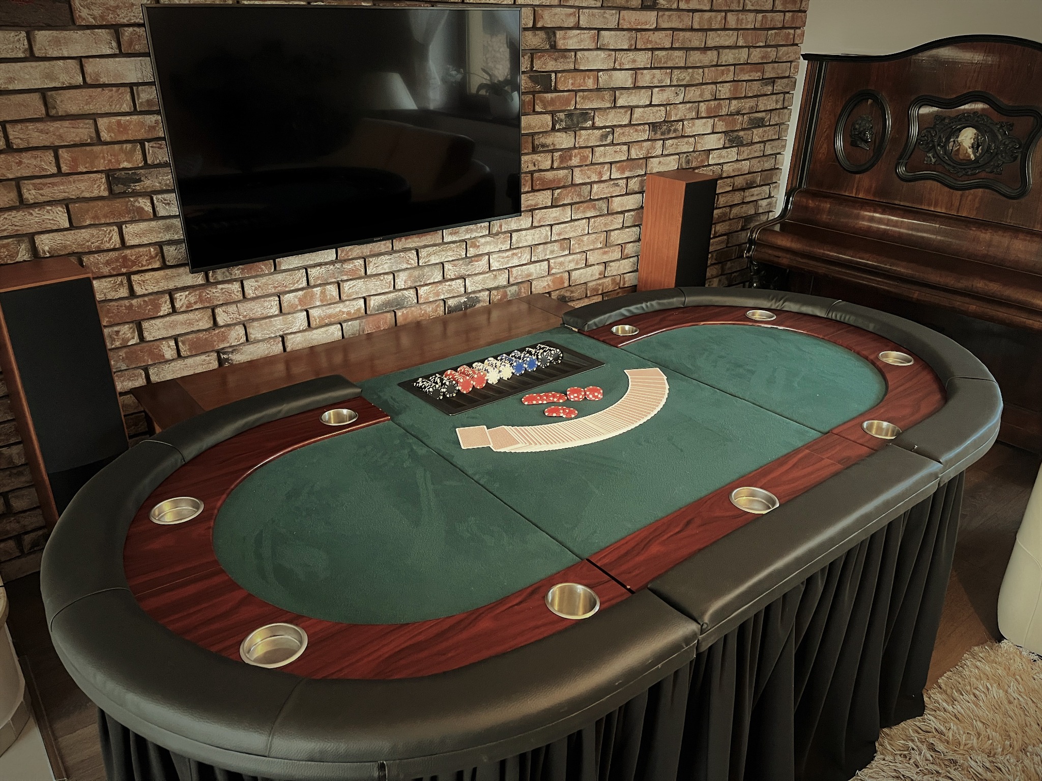 Stół do pokera z zielonym suknem, żetonami i kartami w eleganckim wnętrzu z ceglaną ścianą i fortepianem w tle.