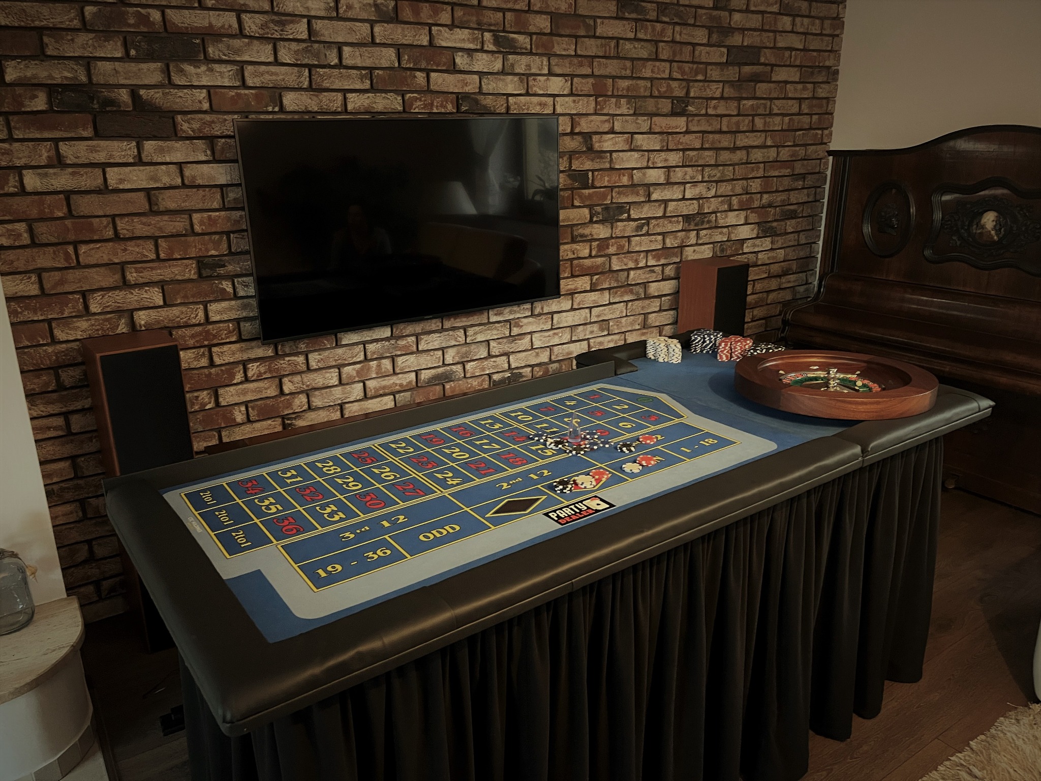 Stół do ruletki z żetonami i kołem ruletki, ustawiony w pomieszczeniu z ceglaną ścianą i telewizorem. Widoczny napis 'Party Dealers'.