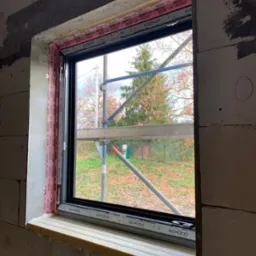 Montaż okna PCV z czarną ramą w surowym murze, zabezpieczone taśmą okienną Schüco, widok na rusztowanie i drzewa w tle.