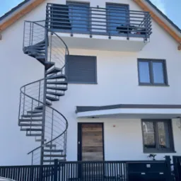 Biały dom z szarą, metalową klatką schodową spiralną prowadzącą na balkon; widoczne szare okna, drzwi z drewnianą okładziną i czarne skrzynki pocztowe.