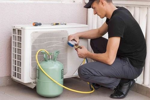 Mężczyzna w czarnej koszulce i czapce kuca przed białą jednostką klimatyzacyjną, podłączając do niej zieloną butlę z czynnikiem chłodniczym za pomocą żółtego węża, narzędzia leżą na górze...