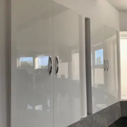 Białe, lakierowane szafki kuchenne z chromowanymi uchwytami, odbijające światło okien i otoczenia, zamontowane nad blatem z ciemnego, nakrapianego kamienia.