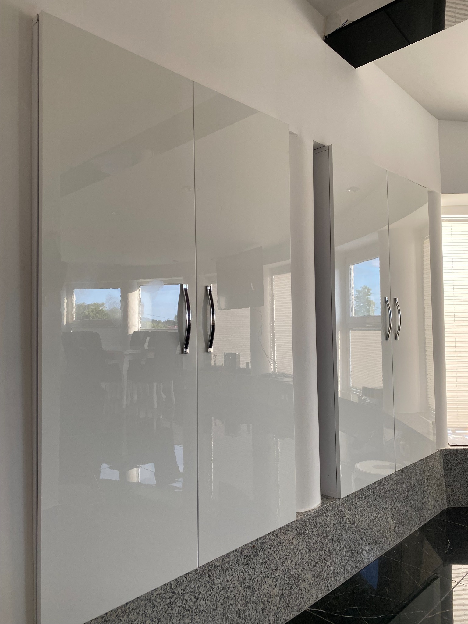 Białe, lakierowane szafki kuchenne z chromowanymi uchwytami, odbijające światło okien i otoczenia, zamontowane nad blatem z ciemnego, nakrapianego kamienia.