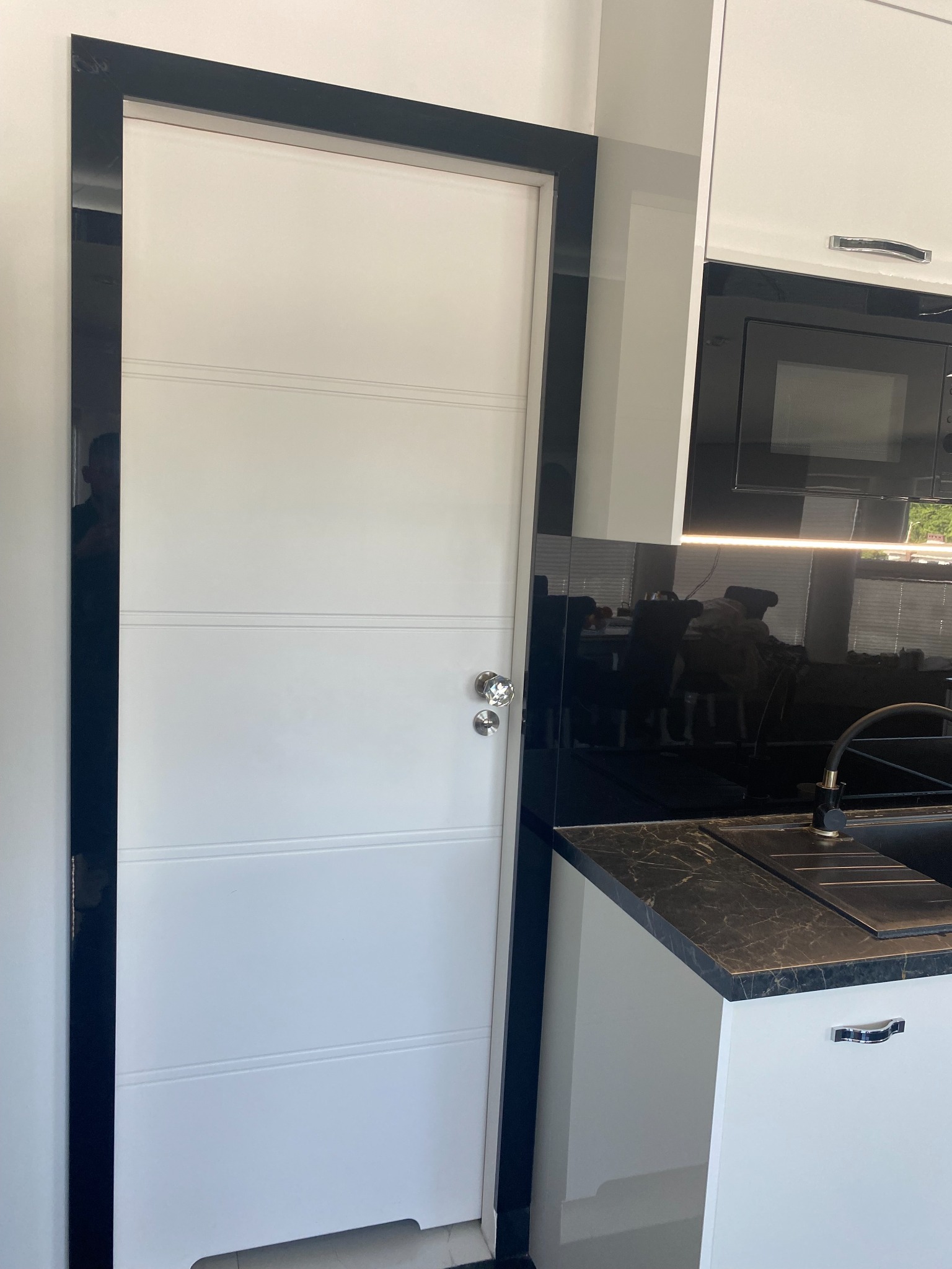 Nowoczesna kuchnia z białymi frontami, czarnym blatem z marmurowym wzorem i czarnym panelem szklanym na ścianie, widoczny fragment drzwi z czarną ramą i kryształową gałką.