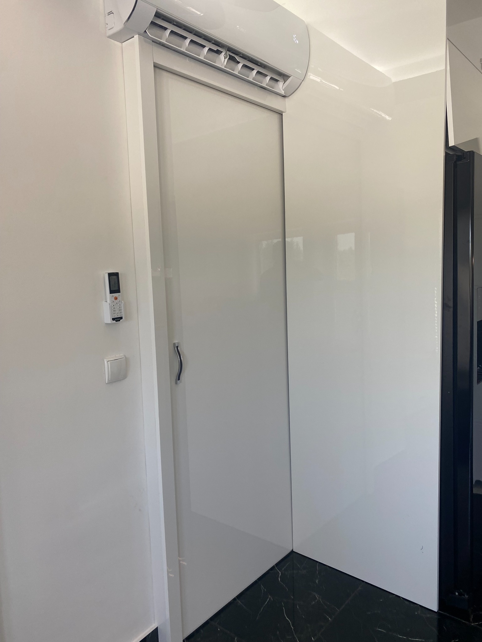 Biała szafa wnękowa z przesuwnymi drzwiami, zamontowana pod klimatyzatorem, z widocznym pilotem i czarną marmurową podłogą.