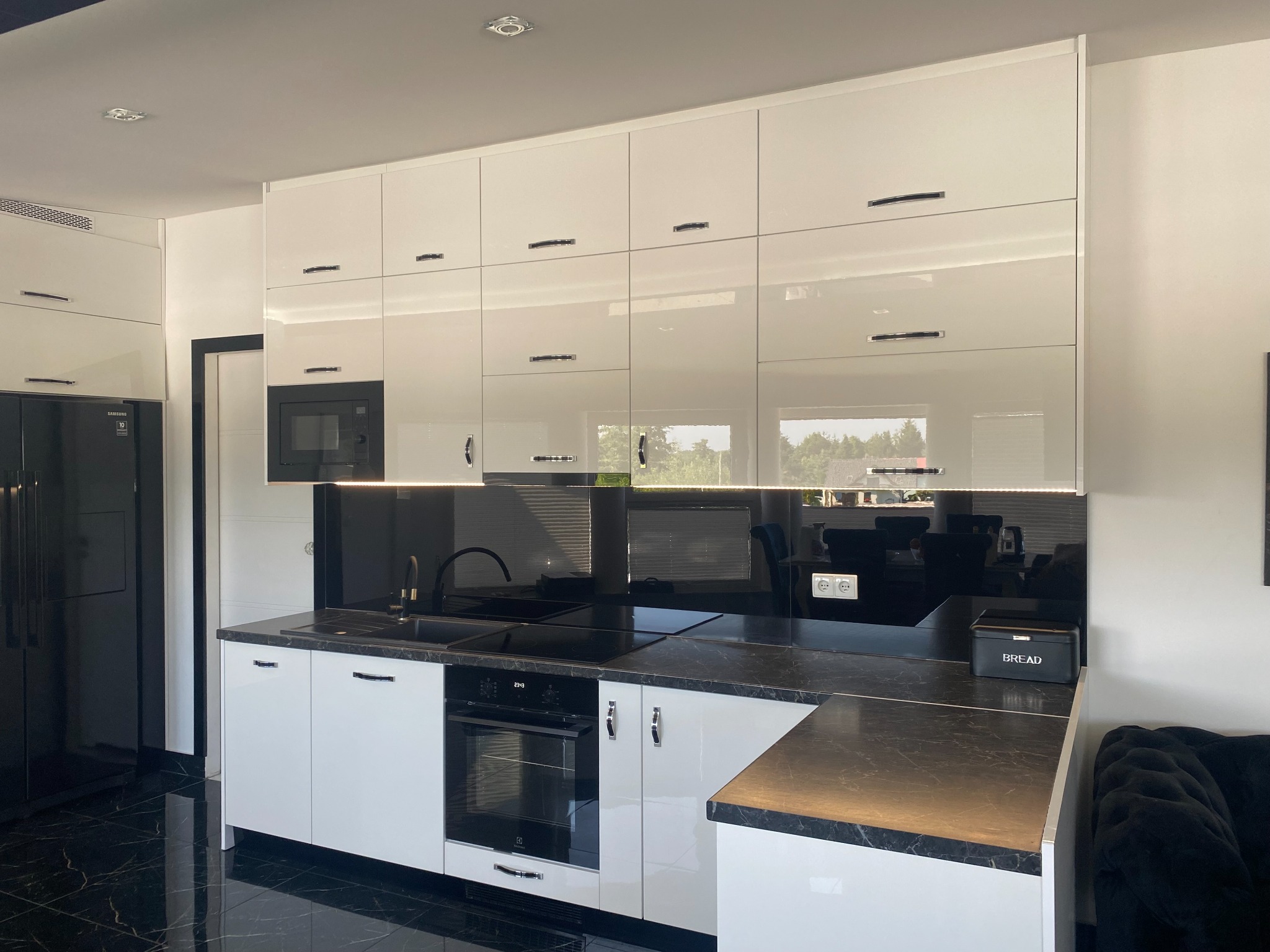 Nowoczesna kuchnia z białymi, lakierowanymi szafkami, czarnym blatem i czarnym szklanym panelem nad blatem, uzupełniona o czarną lodówkę i czarną podłogę z efektem marmuru.