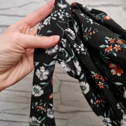 Anastasiya Kazhanouskaya - Dłoń przytrzymuje marszczony rękaw czarnej sukienki w białe i pomarańczowe kwiaty, z wiązaniem z tej samej tkaniny, na tle ściany z białej cegły.