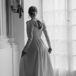 Anastasiya Kazhanouskaya - Kobieta w długiej, białej sukni z wycięciem na plecach ozdobionym koronką, stojąca tyłem w eleganckim wnętrzu z kolumnami i świecznikiem.