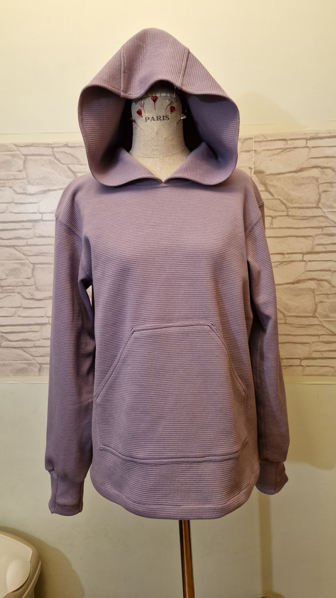 Liliowa bluza z kapturem typu kangurka, uszyta z tkaniny o fakturze wafla, prezentowana na manekinie krawieckim z napisem 'Paris' i szpilkami w kształcie serc, na tle ściany imitującej cegłę.