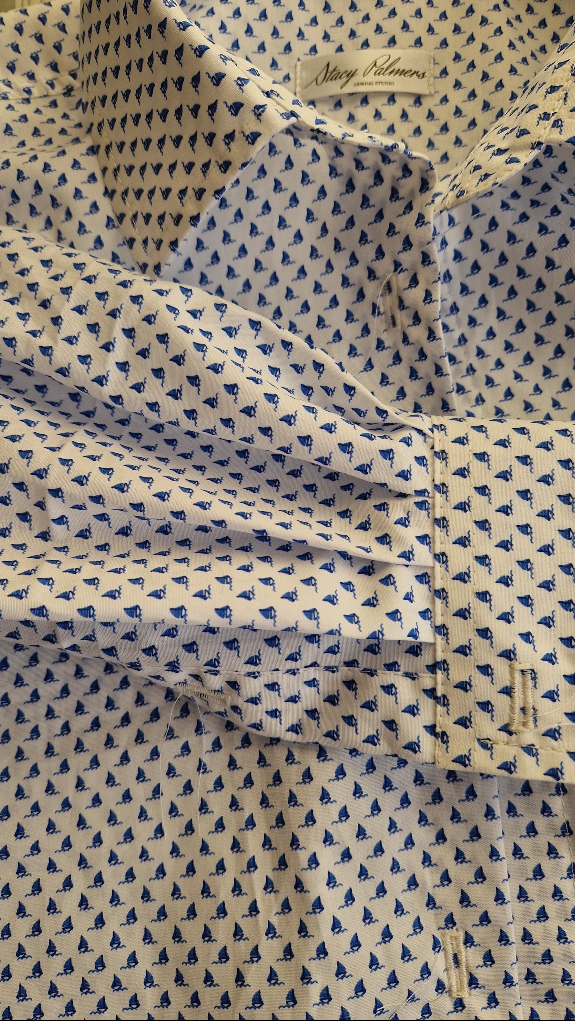 Fragment białej koszuli męskiej z niebieskim wzorem żaglówek, widoczny mankiet i kołnierzyk, bliski kadr na tkaninę.