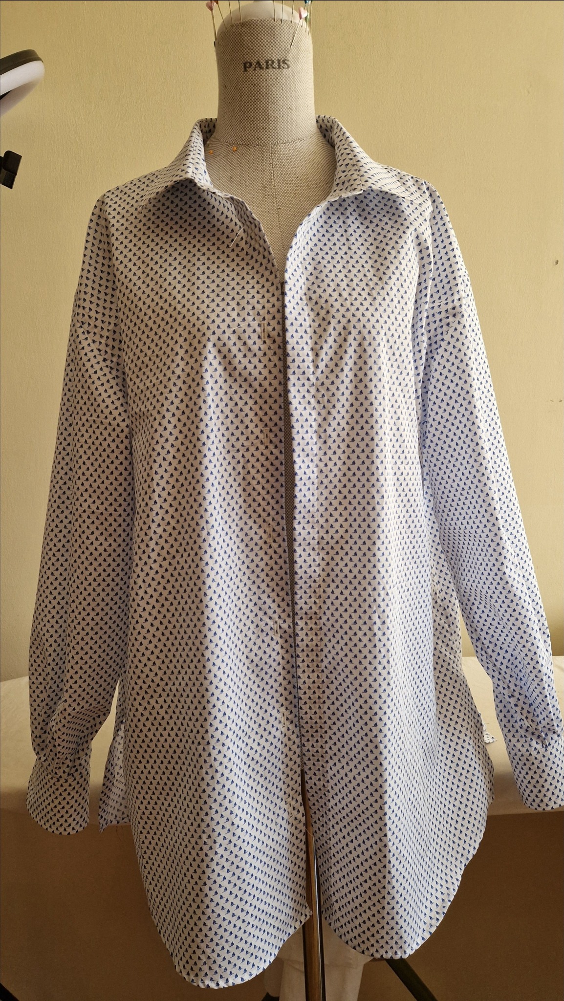 Niedokończona biała koszula męska w drobne niebieskie żaglówki na manekinie krawieckim z napisem PARIS, widoczne szpilki w kołnierzyku.