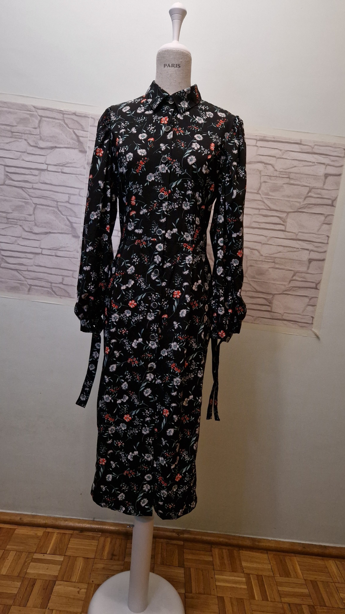 Czarna sukienka koszulowa w drobne kwiaty, z długimi rękawami i wiązaniem w pasie, prezentowana na manekinie w jasnym pomieszczeniu z tłem imitującym cegły.